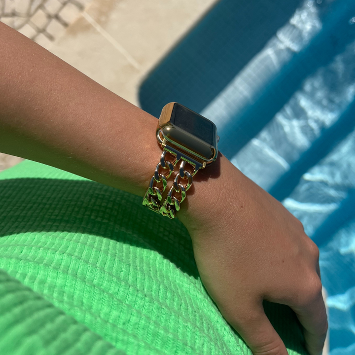 A person wearing a gold-chain wristwatch with a rectangular face is resting their arm near a pool. They are dressed in a bright green textured garment and have an Apple Watch outfitted with the Coconut Lane 2-in-1 Metallic Apple Watch Case & Built-In Screen Protector in Gold. The background features part of the pool's edge and the surrounding tiles.