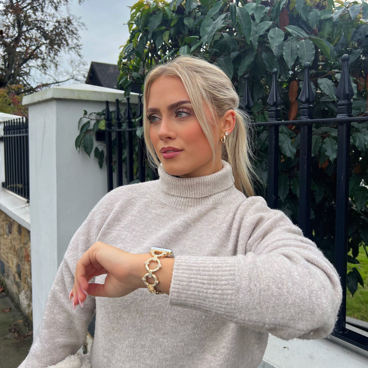 A person with blonde hair in a ponytail is standing outdoors near a black iron fence and green bushes. They are wearing a light-colored turtleneck sweater, gold jewelry, and an Apple Watch with a Diamante Bracelet Apple Watch Strap by Coconut Lane, looking to the side with one hand resting on their wrist.