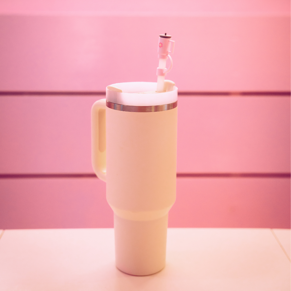 A Mini Pink Tumbler Straw Cover from Coconut Lane is placed on a pink and white surface. The background features blurred pink horizontal panels.