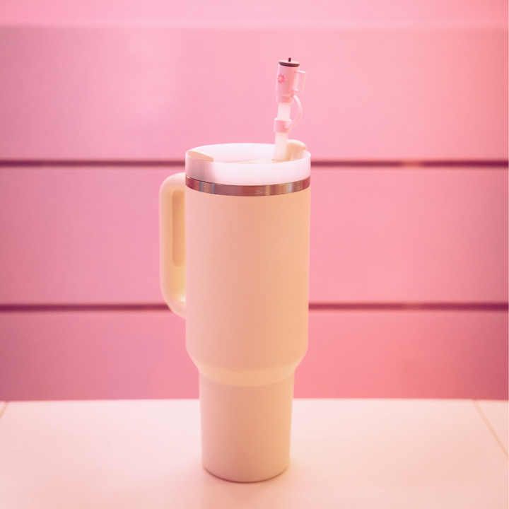 A Mini Pink Tumbler Straw Cover from Coconut Lane is placed on a pink and white surface. The background features blurred pink horizontal panels.