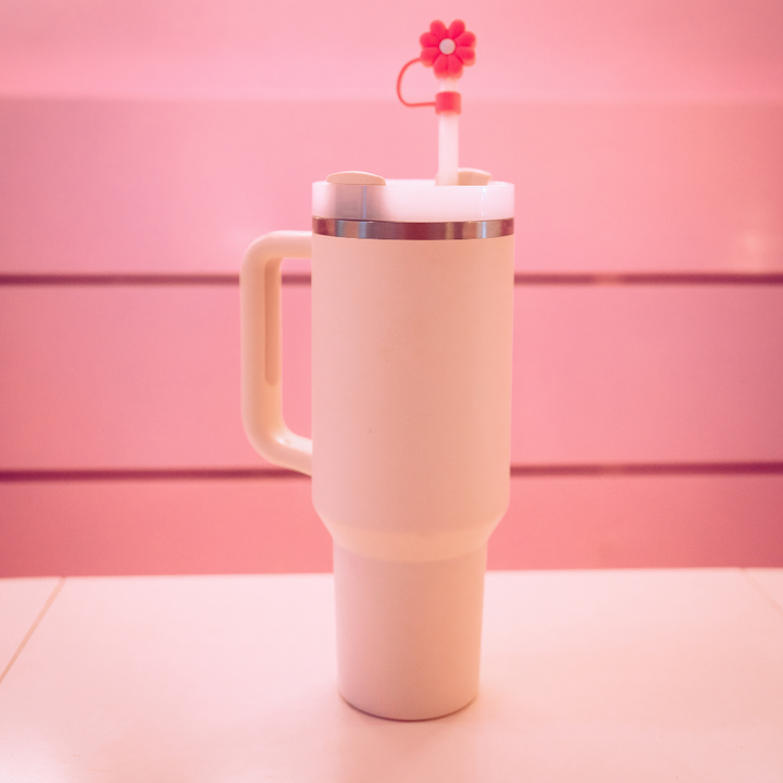 A large white insulated tumbler with a handle and a stainless steel rim sits on a pink surface, against a pink background. The tumbler is topped with a Coconut Lane Pink & White Daisy Straw Cover placed on its lid's straw.