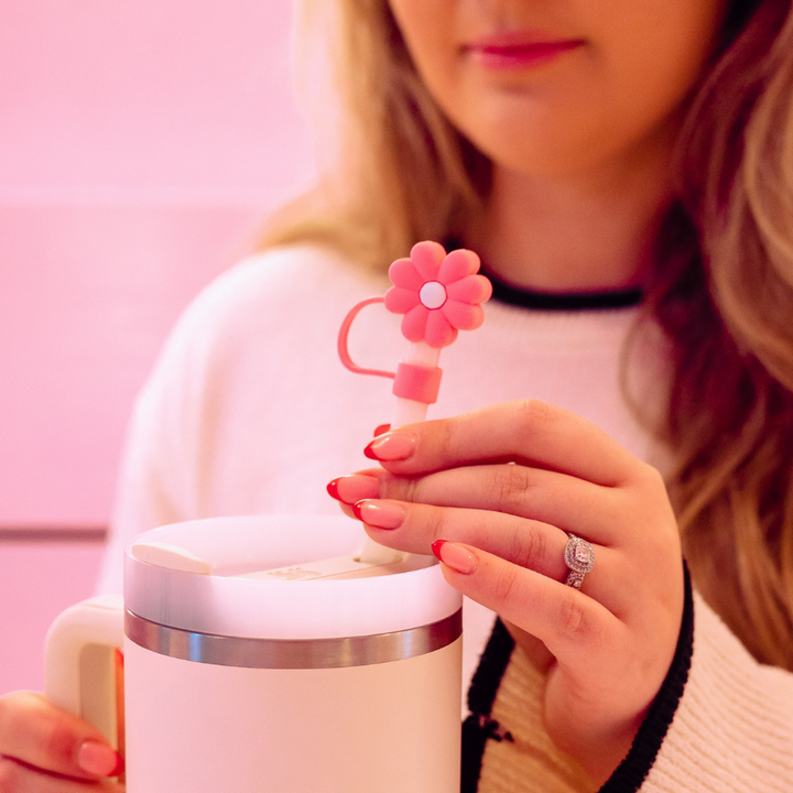 A woman with long hair is holding a white container with a metallic rim. She is inserting the Coconut Lane Pink & White Daisy Straw Cover into the container. She is wearing a ring on her left hand and has manicured nails painted in soft pink. The background is a gradient of pink hues.