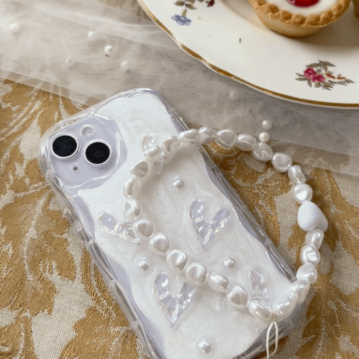 A white iPhone with a Coconut Lane Pearly Hearts Phone Case - Clear, showcasing pearl-like accents and transparent heart shapes, rests on an ornate gold-patterned tablecloth. A white bracelet with pearl-like beads lies atop the phone. In the background, there is a floral-patterned dessert plate filled with pastries.