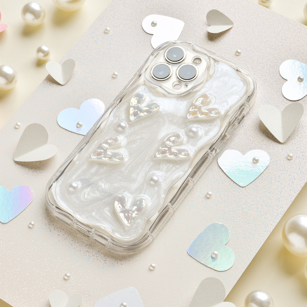 Introducing the Pearly Hearts Phone Case - Clear from Coconut Lane for the iPhone 15. This extra protective case showcases raised, shiny heart designs against a pearlescent, swirled background. It elegantly complements any look while your phone rests on a beige, glittery surface adorned with metallic heart confetti and pearl-like beads. Combining style and safety seamlessly, it's the perfect blend of functionality and glamour.