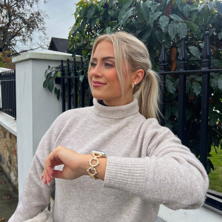 A woman with blonde hair tied back in a ponytail is standing outdoors in front of a fence and greenery. She is wearing a light gray turtleneck sweater, accessorized with Coconut Lane's Diamante Bracelet Apple Watch Strap compatible with all Apple Watch models, and she is smiling while looking to the side.