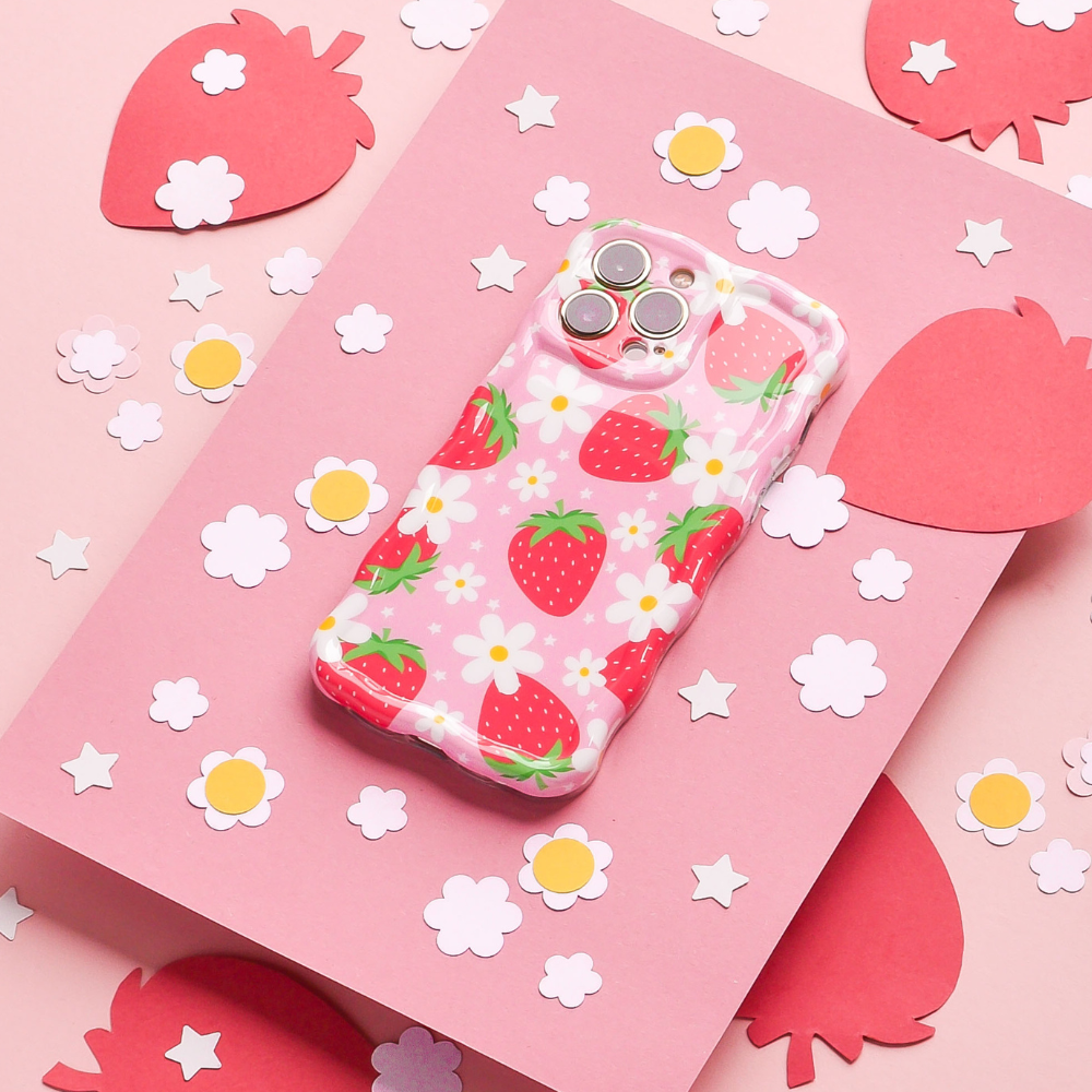 A Coconut Lane Wavy Phone Case - Berry Cute Strawberry with a delightful strawberry pattern and white flowers rests on a pink surface, surrounded by cut-out paper shapes of strawberries and flowers. The background includes larger pink strawberry shapes and smaller white flower shapes scattered around.
