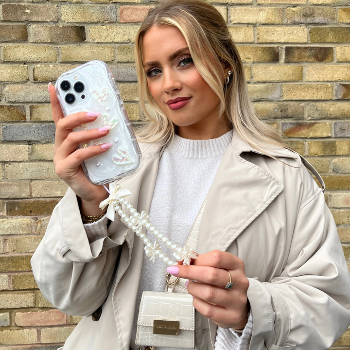 A woman with long blonde hair is holding a smartphone adorned with Coconut Lane's Pearly Hearts Phone Case - Clear, featuring a beaded strap. She is wearing a beige trench coat over a white sweater and has a small white handbag hanging from her shoulder. Standing in front of a brick wall, she poses confidently for the camera.