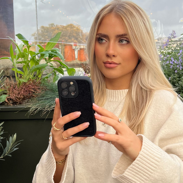 A woman with long blonde hair and a light-colored sweater is sitting outdoors, holding a smartphone with the Cosy Teddy Phone Case - Black from Coconut Lane. She appears focused, with greenery and potted plants in the background. She's wearing several rings and a bracelet, with a calm expression on her face.