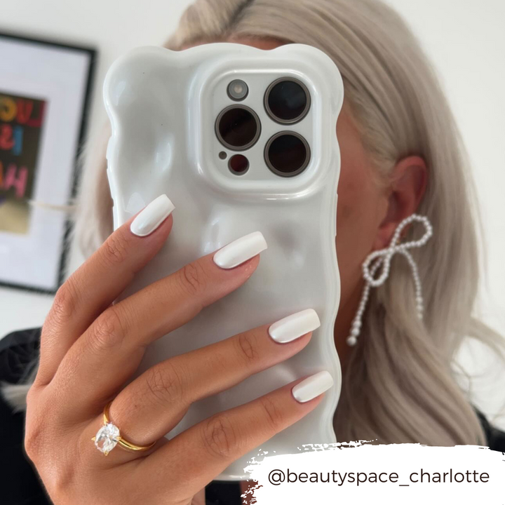 A person with platinum blonde hair is holding a Coconut Lane 3D Pearl Phone Case - White, featuring a wavy design and pearl white effect, obscuring most of their face. They have white manicured nails, a diamond ring on their ring finger, and are wearing bow-shaped earrings. The Instagram handle @beautyspace_charlotte is visible.