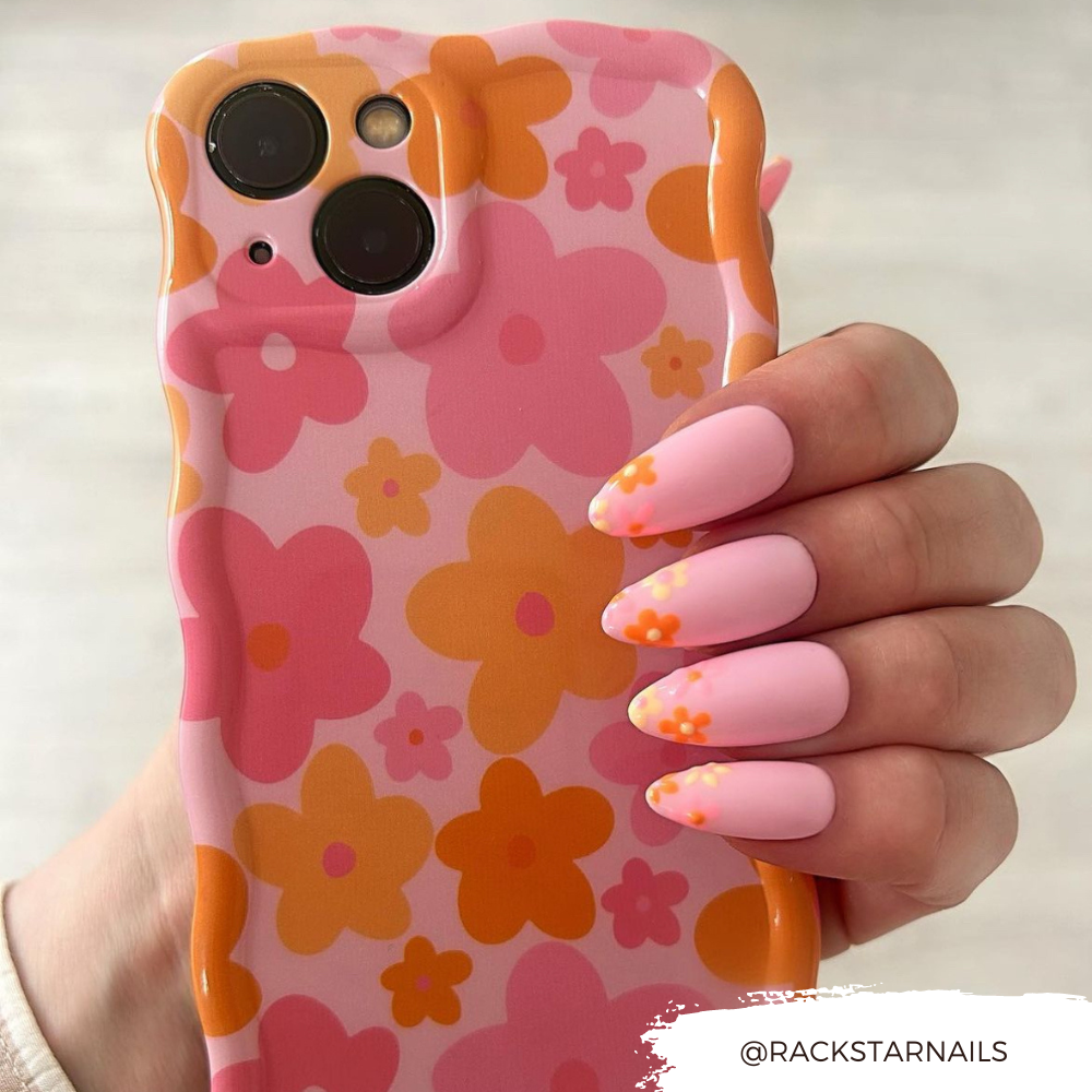 A hand with pastel pink nails featuring orange floral designs holds a Coconut Lane Wavy Phone Case - Pink & Orange Bloom. The user's handle @RACKSTARNails is displayed in the bottom right corner.
