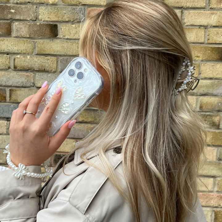 A woman with long, wavy blonde hair holds her iPhone15 adorned with the Pearly Hearts Phone Case - Clear by Coconut Lane to her ear. She is wearing a beige trench coat and a pearl bracelet. Her hair is accessorized with a decorative hair clip. The background features a brick wall, adding to the chic ambiance.