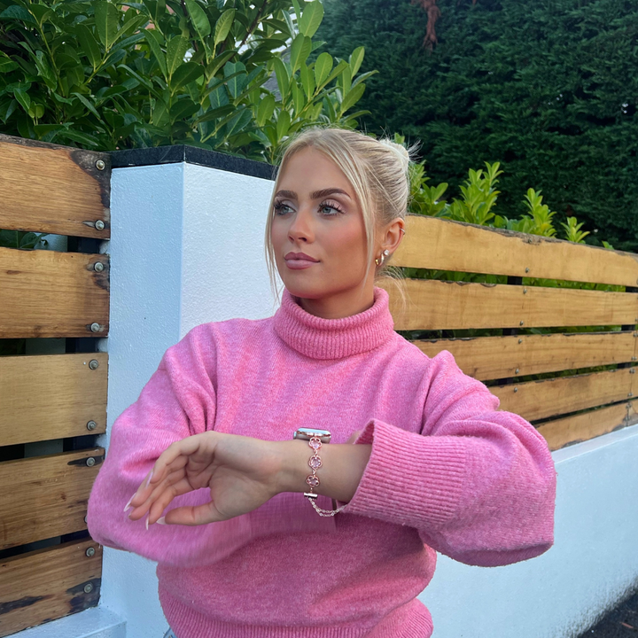 A woman with blonde hair tied back in a ponytail is wearing a pink turtleneck sweater, sitting outdoors in front of a wooden fence and green foliage. She is looking off to the side, showcasing her stylish rose gold jewelry and the Chain Bracelet Apple Watch Strap - Rose Gold by Coconut Lane.