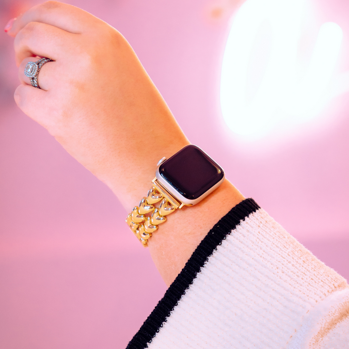 A person flaunts a Hearts Chain Apple Watch Strap - Gold by Coconut Lane on their wrist, complementing the diamond ring on their finger. The soft pink background with blurred white lighting adds elegance, while the sleeve of their white top with black trim is partially visible.