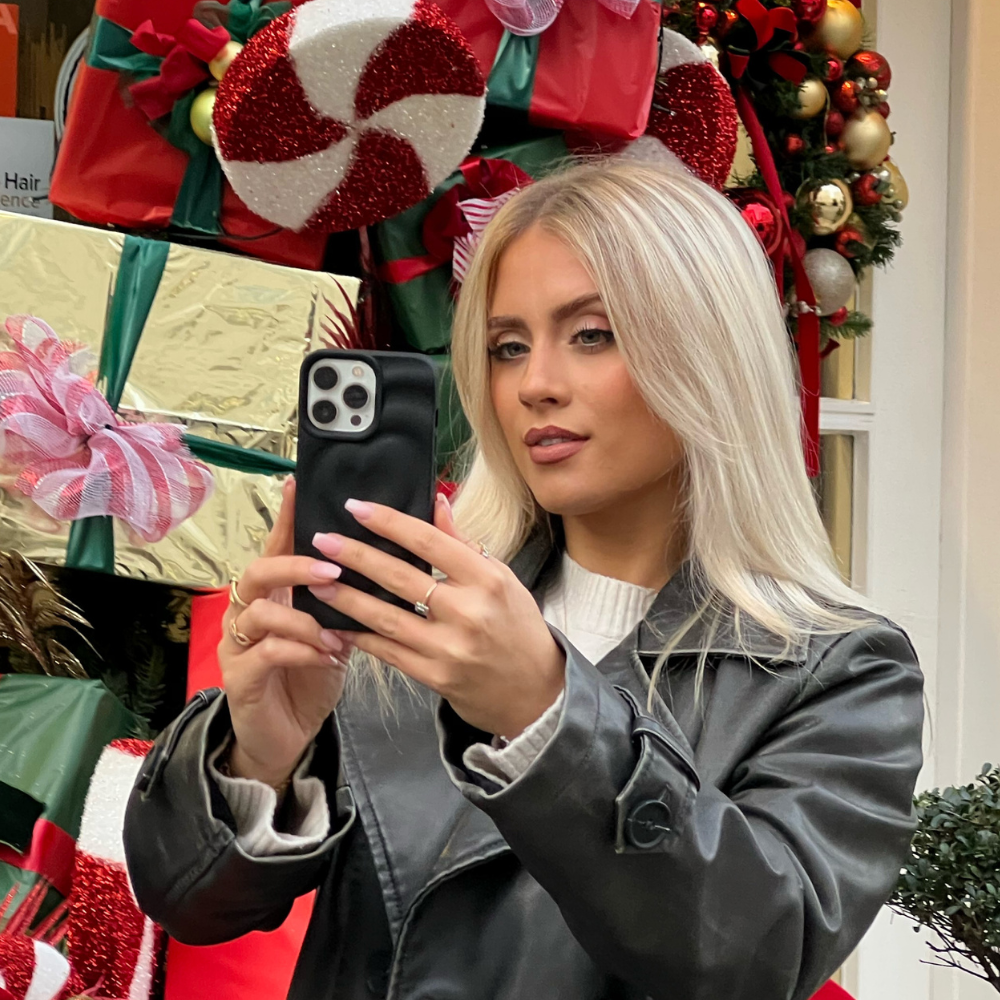 A blonde woman in a black leather jacket takes a selfie with her Coconut Lane Melting Heart Phone Case - Matte Black. She stands in front of festive decorations, including large wrapped presents and candy canes adorned with red bows and ribbons. The background suggests a holiday theme with greenery and ornaments.