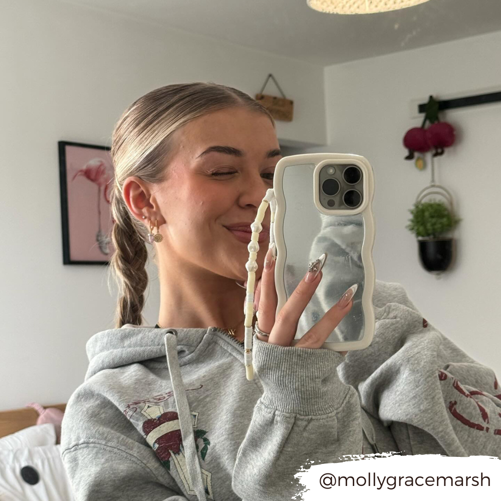 A person in a grey hoodie takes a mirror selfie using a phone protected by the stylish Curvy Mirror Phone Case - Cream from Coconut Lane. They are winking and smiling, with their hair styled in a braid. The room behind them includes a bed, wall art, and hanging decorations. The username "@mollygracemarsh" is visible.