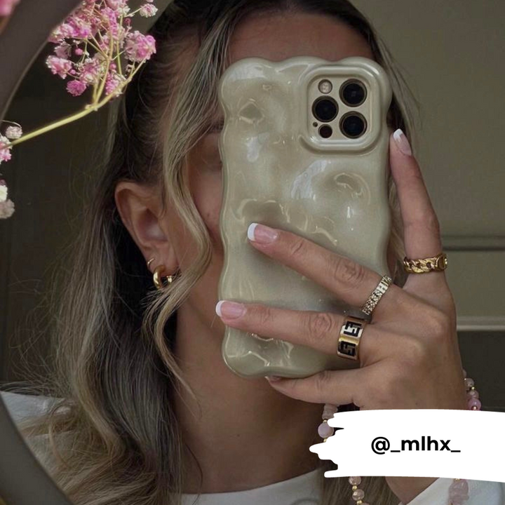 A person with wavy hair holds an iPhone 15 featuring the Coconut Lane 3D Pearl Phone Case - Gold in front of their face. They wear gold hoop earrings, a gold ring, and have manicured nails. There are pink flowers visible in the upper left corner. The Instagram handle @_mlhx_ is displayed at the bottom.