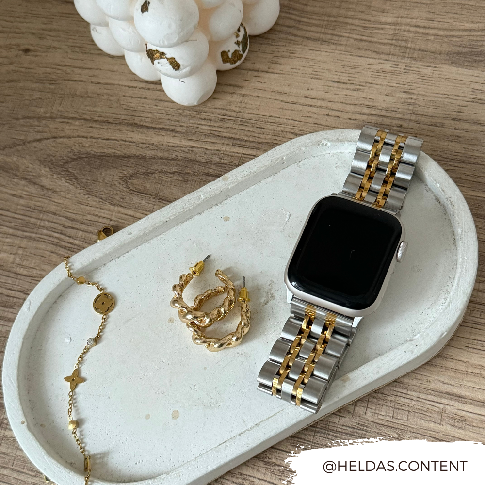 A Stainless Steel Apple Watch Strap - Silver & Gold by Coconut Lane with an adjustable strap rests on a decorative white tray, accompanied by gold earrings and a bracelet with a star charm. The tray sits on a wooden surface, featuring the Instagram handle @heldas.content in the bottom right corner.