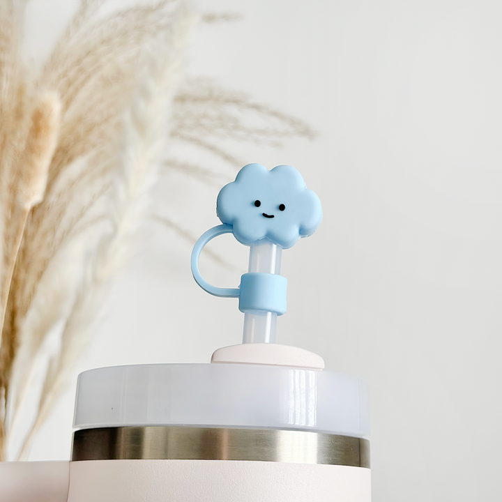 The close-up image showcases the Blue Cloud Straw Cover by Coconut Lane, a light blue, cloud-shaped straw topper made of high-quality silicone with a cheerful smiley face. It sits atop a cup with a transparent lid accented by a metal band. The background features softly-focused, wheat-like plants set against a neutral pastel backdrop.