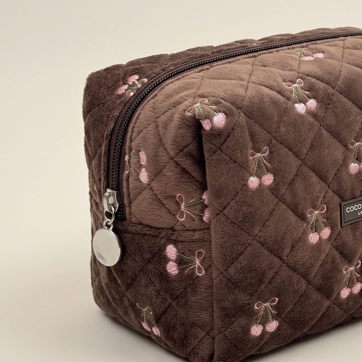 Close-up of a brown quilted velvet makeup bag with pink embroidered cherries and a round silver zip pull.
