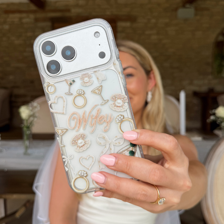 Bride holding a phone case with rings, hearts, cocktails, bows, pearls and the word 'Wifey'