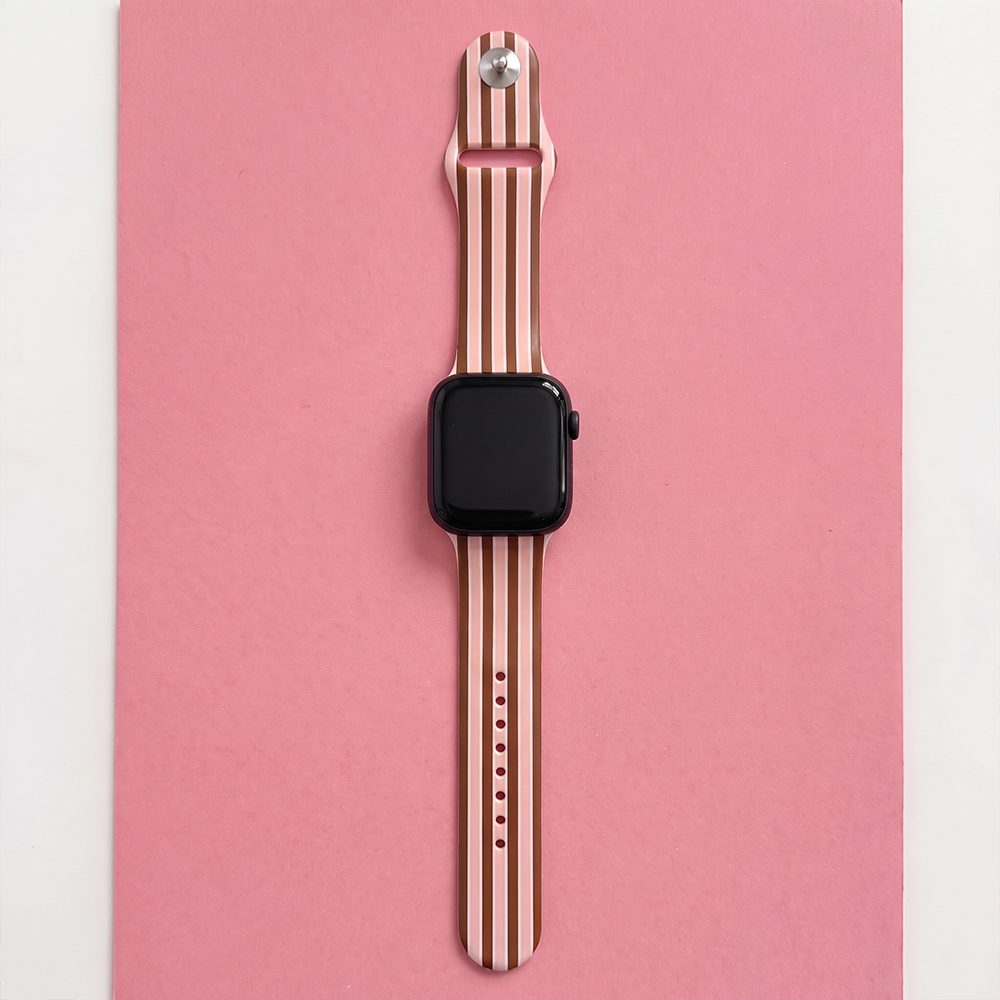 Overhead flatlay of the Cocoa & Pink Stripe Apple Watch strap, featuring vertical brown, pink and cream stripes, adjustable holes and a black Apple Watch on a pink background.