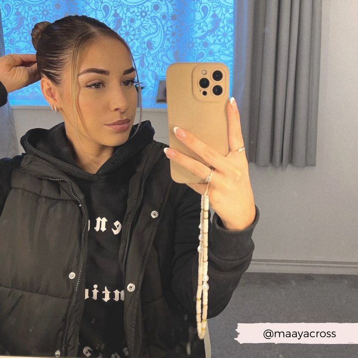 A woman with her hair in a bun takes a mirror selfie with her iPhone, adorned with the stylish Coconut Lane Beaded Phone Strap in Nude & Pearl. She is wearing a black hoodie and a black puffer vest. The background shows a window with decorative curtain and cream-colored drapes. A username "@maayacross" is displayed in the bottom right corner.