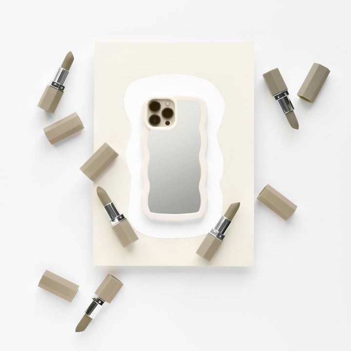 A stylish iPhone adorned with a sleek Curvy Mirror Phone Case in Cream from Coconut Lane lies on a white and beige background, surrounded by six lipsticks with beige casings. The composition seamlessly blends tech and beauty products, creating a minimalist and elegant visual.