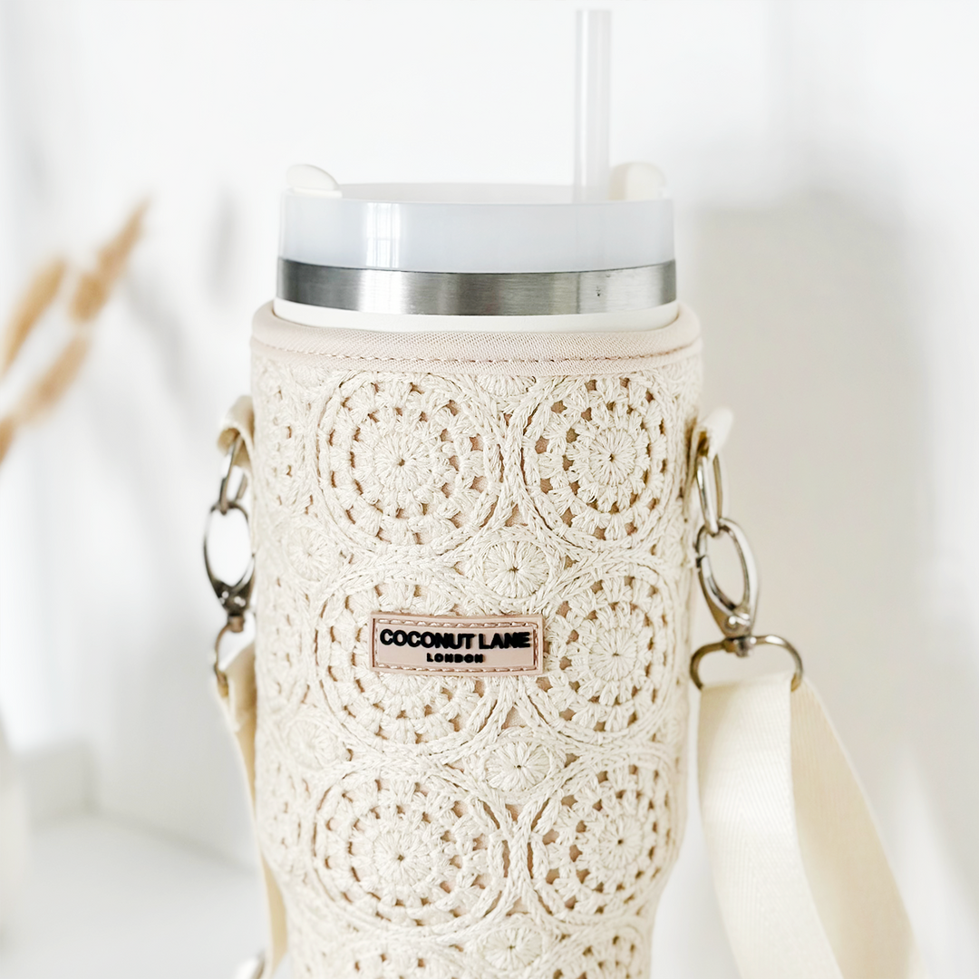 A close-up of a white tumbler protected by the Tumbler Carry Case - Festival Crochet by Coconut Lane, featuring a textured, intricately crocheted sleeve with floral patterns. The tumbler has a white lid with a transparent straw and beige shoulder straps attached on both sides, making it the perfect travel accessory. A label on the sleeve reads "Coconut Lane London.