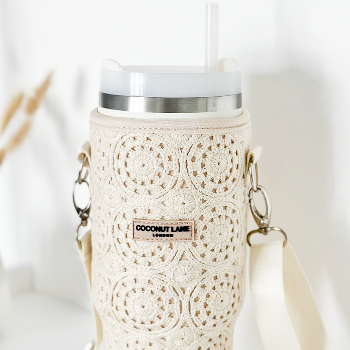 A close-up of a white tumbler protected by the Tumbler Carry Case - Festival Crochet by Coconut Lane, featuring a textured, intricately crocheted sleeve with floral patterns. The tumbler has a white lid with a transparent straw and beige shoulder straps attached on both sides, making it the perfect travel accessory. A label on the sleeve reads "Coconut Lane London.