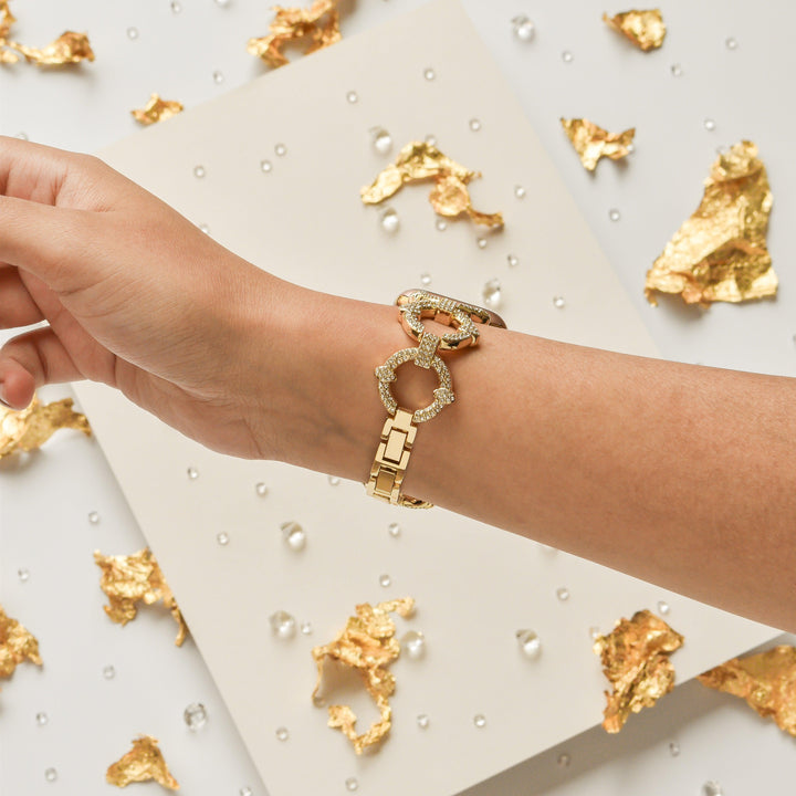 A hand wearing a gold bracelet adorned with interlocking circular designs set with diamonds. The backdrop consists of scattered gold leaf pieces and small clear beads, all placed on a white surface, next to the chic Diamante Bracelet Apple Watch Strap by Coconut Lane.