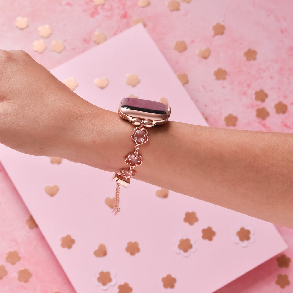 A hand showcasing a sleek Rose Gold Flower Bracelet Apple Watch Strap by Coconut Lane, adorned with a matching rose gold charm bracelet featuring pink and gold accents. The background consists of a pink surface scattered with gold floral and heart-shaped confetti, creating an elegant and coordinated look.
