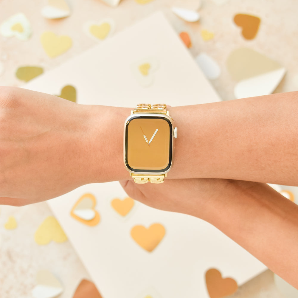 Close-up of a person wearing a gold smartwatch with the Coconut Lane Hearts Chain Apple Watch Strap - Gold. The background is adorned with various gold and cream-colored heart shapes. The person's hand is resting on what appears to be another arm or hand, elegantly showcasing the watch.