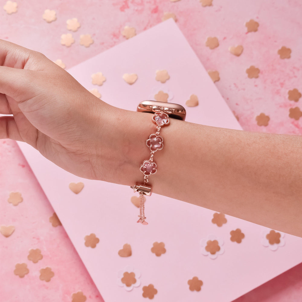 A hand wearing the Coconut Lane Rose Gold Flower Bracelet Apple Watch Strap with pink and rose gold heart-shaped charms is shown against a pink background with gold and white flower-shaped confetti.