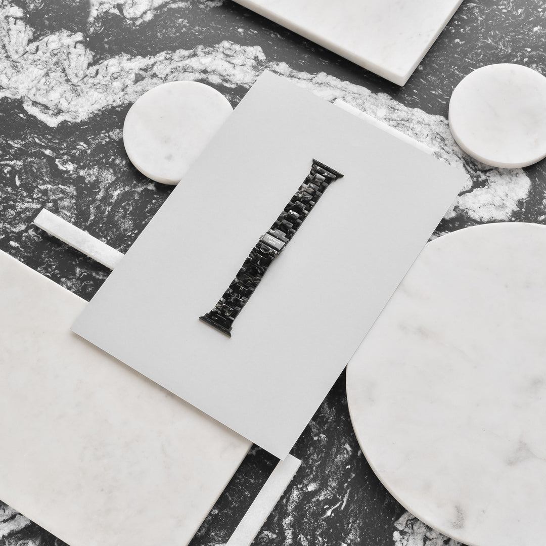 A Luxe Black Marble Apple Watch Strap from Coconut Lane is displayed on a white rectangular card, laid on a marble surface with various round and rectangular marble pieces around it. The watch strap is centered and stands out against the contrasting white and grey marbled background.