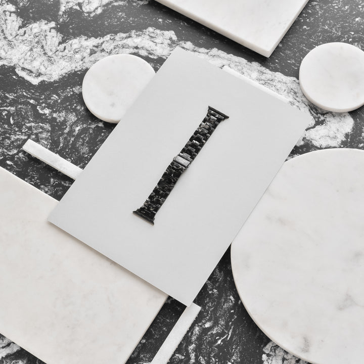 A Luxe Black Marble Apple Watch Strap from Coconut Lane is displayed on a white rectangular card, laid on a marble surface with various round and rectangular marble pieces around it. The watch strap is centered and stands out against the contrasting white and grey marbled background.