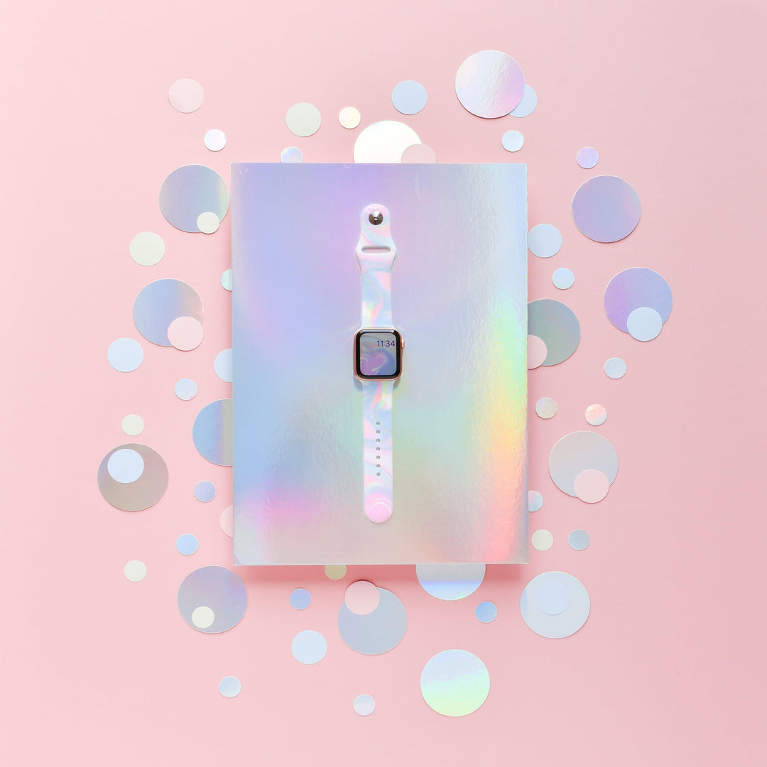 A white smartwatch with a square screen is centered on a holographic, iridescent background. The Holo Bubble Apple Watch Strap by Coconut Lane stands out, complementing the backdrop's mix of circular cutouts in various sizes. The entire scene is enveloped in a soft, pastel pink tone.