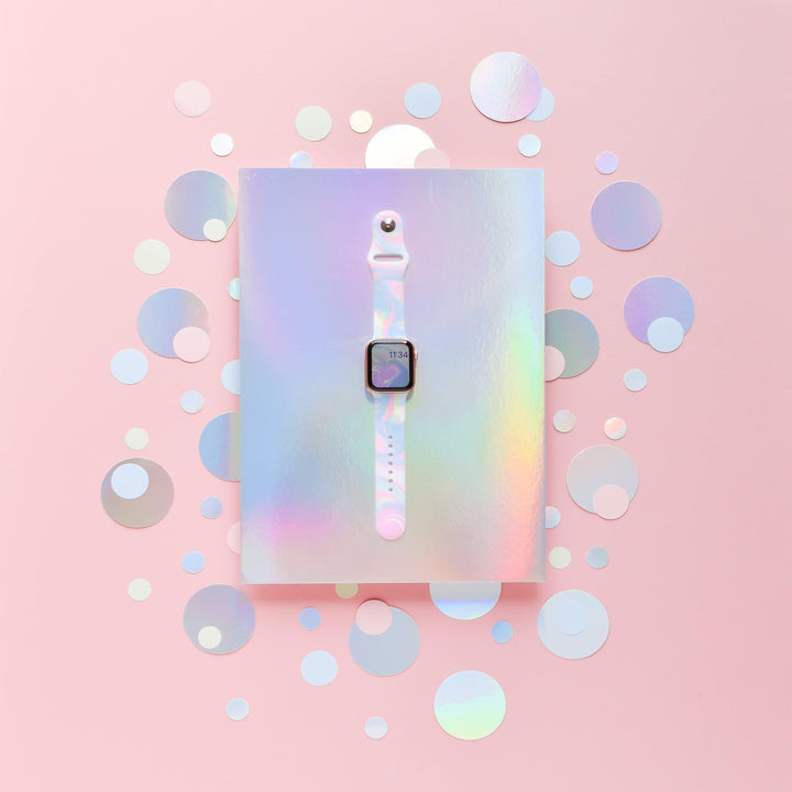A white smartwatch with a square screen is centered on a holographic, iridescent background. The Holo Bubble Apple Watch Strap by Coconut Lane stands out, complementing the backdrop's mix of circular cutouts in various sizes. The entire scene is enveloped in a soft, pastel pink tone.