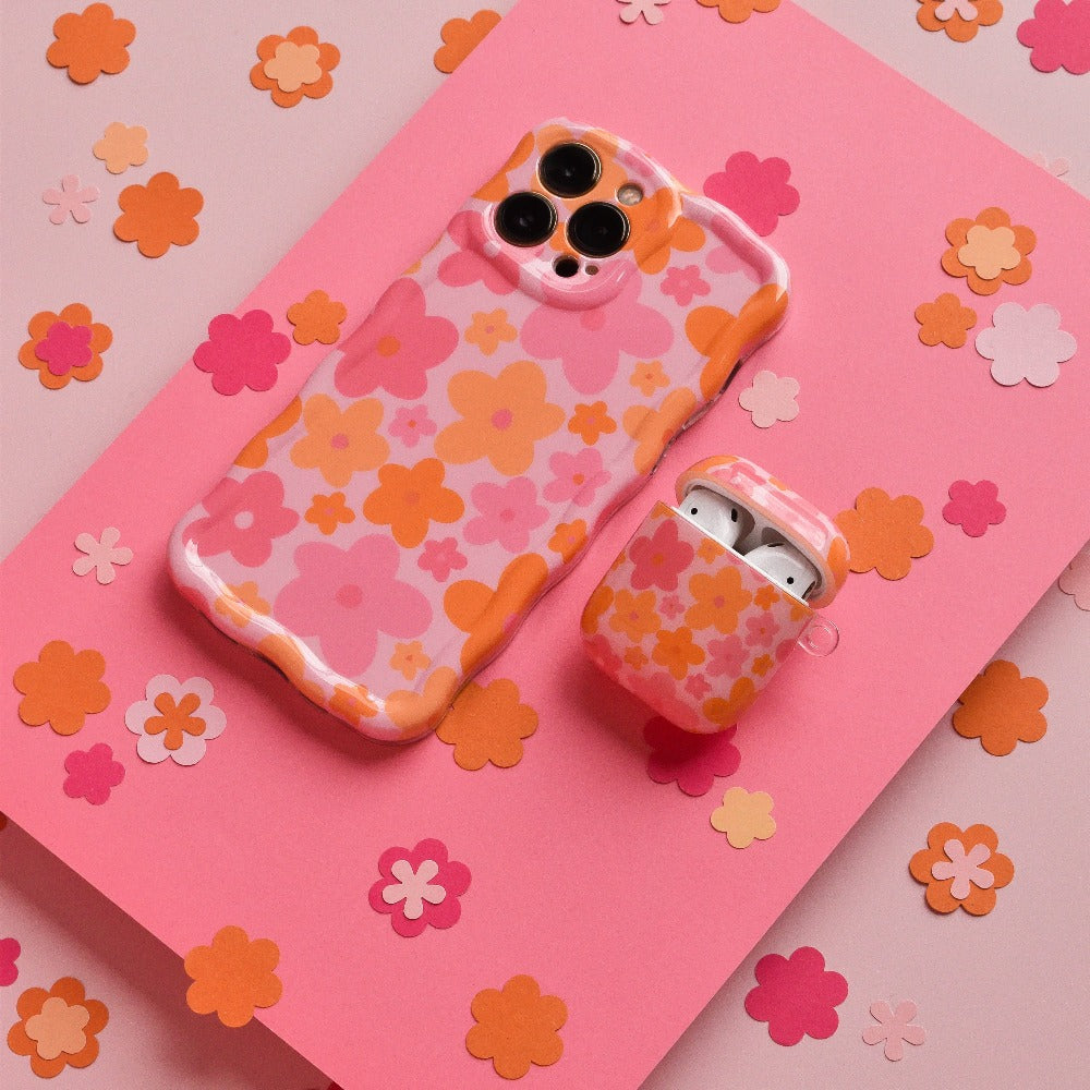 A Wavy Phone Case - Pink & Orange Bloom by Coconut Lane and an AirPods case, both covered in matching floral designs with pink, orange, and peach-colored flowers, are placed on a pink background scattered with small paper flowers in similar colors. This extra protective duo adds a charming touch to your tech essentials.