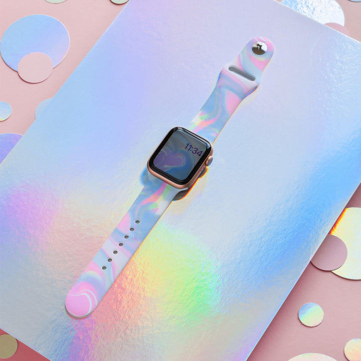 A smart watch featuring Coconut Lane's Holo Bubble Apple Watch Strap, adorned with a vibrant pastel marble pattern, is showcased on a reflective, iridescent surface. The watch screen indicates the time as 11:34. Circular holographic confetti pieces are scattered around the watch on a pink background.