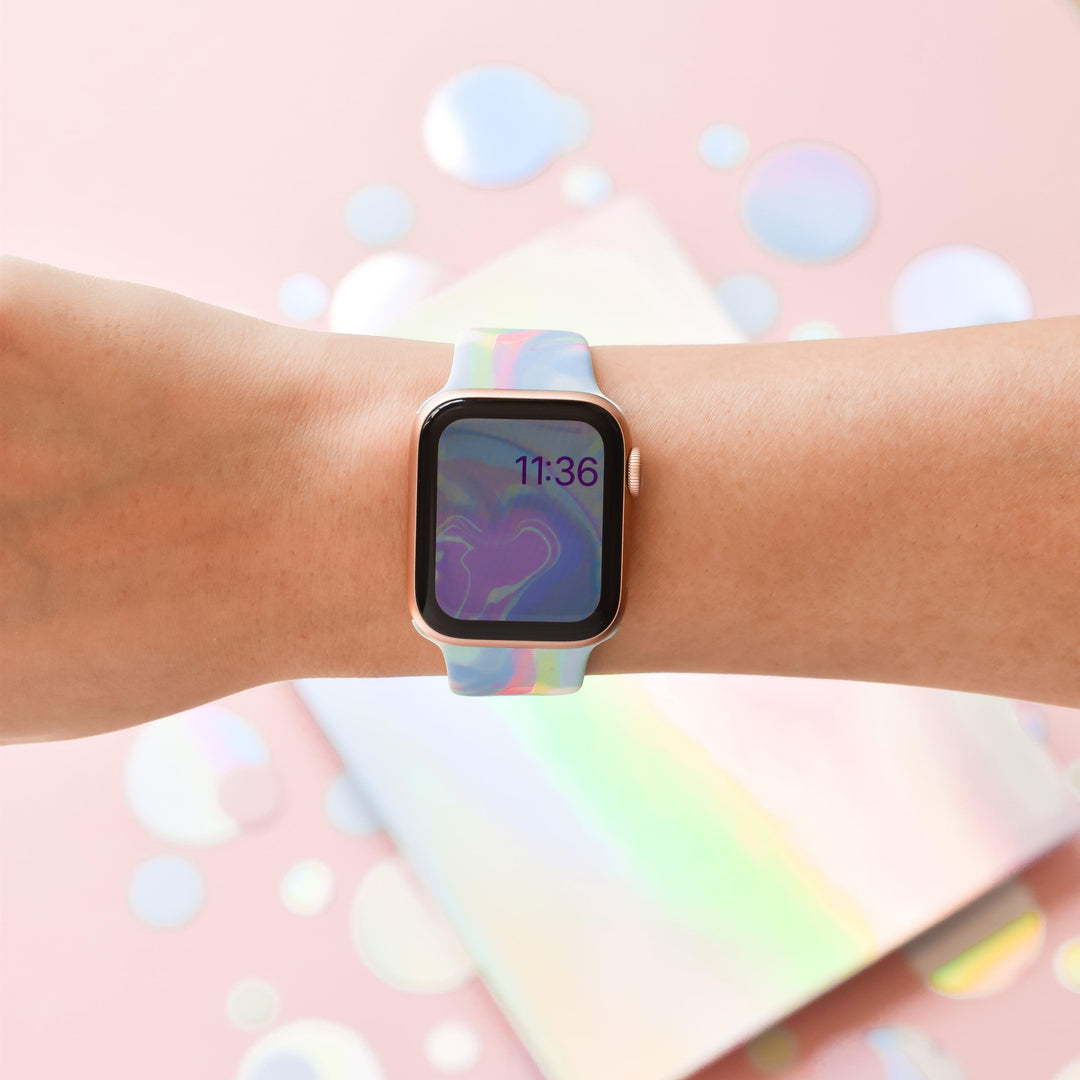 A person wears the Coconut Lane Holo Bubble Apple Watch Strap, featuring a colorful, holographic iridescent design on their wrist. The watch face displays the time as 11:36 against a wavy, abstract background. The backdrop is a pastel pink surface with reflective iridescent circles scattered around.