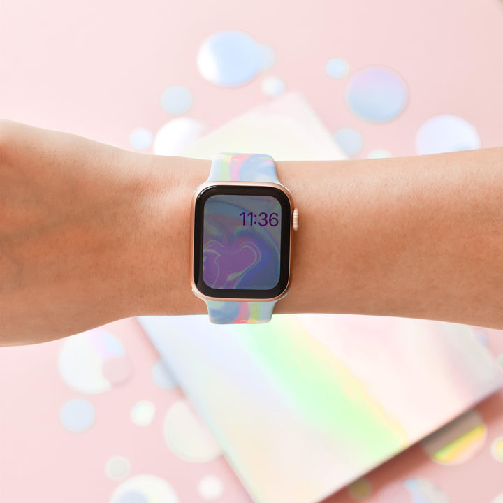 A person wears the Coconut Lane Holo Bubble Apple Watch Strap, featuring a colorful, holographic iridescent design on their wrist. The watch face displays the time as 11:36 against a wavy, abstract background. The backdrop is a pastel pink surface with reflective iridescent circles scattered around.