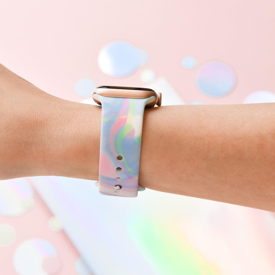 A close-up image of a person's arm wearing an Apple Watch paired with the Coconut Lane Holo Bubble Apple Watch Strap, showcasing its multi-colored, pastel gradient. The background is a soft pink with blurred circular reflections that create a dreamy, abstract atmosphere, perfectly accentuating the strap's holographic iridescent design.