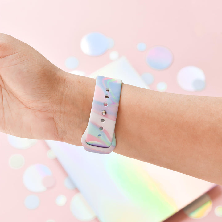 A hand is shown wearing Coconut Lane's pastel-colored, marbled Holo Bubble Apple Watch Strap against a pink background adorned with scattered iridescent circular sequins. The person is holding their wrist up, prominently showcasing the strap's holographic iridescent design.