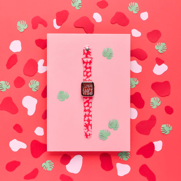 A smartwatch with the Red & Pink Leopard Apple Watch Strap from Coconut Lane is displayed on a pink surface. The background is filled with red, pink, and white abstract shapes and a few green leaf cutouts, creating a vibrant and colorful setting.