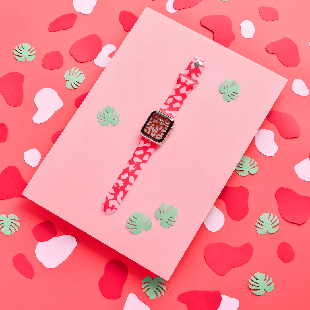 A Coconut Lane Red & Pink Leopard Apple Watch Strap is showcased against a pink paper background. The display is enhanced with green and pink abstract shapes and small paper leaves, creating a whimsical and colorful setup.