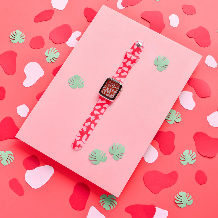 A Coconut Lane Red & Pink Leopard Apple Watch Strap is showcased against a pink paper background. The display is enhanced with green and pink abstract shapes and small paper leaves, creating a whimsical and colorful setup.