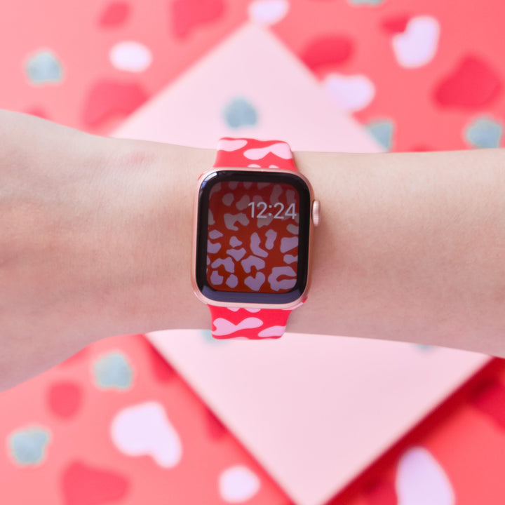 A close-up view of a wrist wearing a Coconut Lane smartwatch with the Red & Pink Leopard Apple Watch Strap and matching display screen showing the time 12:24. The background features scattered heart shapes in various shades of red, pink, and white.