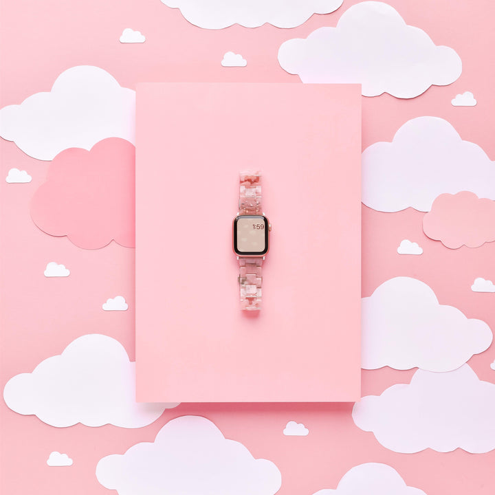 Displayed on a pink rectangular sheet, the "Luxe Pink Clouds Apple Watch Strap" by Coconut Lane showcases its girly and cute design. The background features whimsical white and light pink clouds, enhancing the dreamy aesthetic. The smartwatch displaying a partial time of "1:50" pairs perfectly with this charming accessory, adding flair to any outfit.