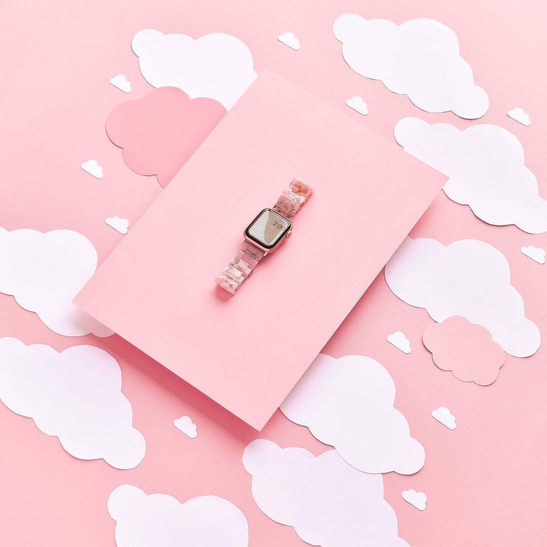 A smartwatch featuring the Luxe Pink Clouds Apple Watch Strap by Coconut Lane is placed on a pink rectangular block. The background consists of off-white and pink cloud cutouts, creating a dreamy, whimsical aesthetic.