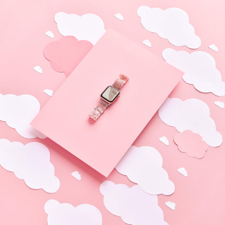 A smartwatch featuring the Luxe Pink Clouds Apple Watch Strap by Coconut Lane is placed on a pink rectangular block. The background consists of off-white and pink cloud cutouts, creating a dreamy, whimsical aesthetic.